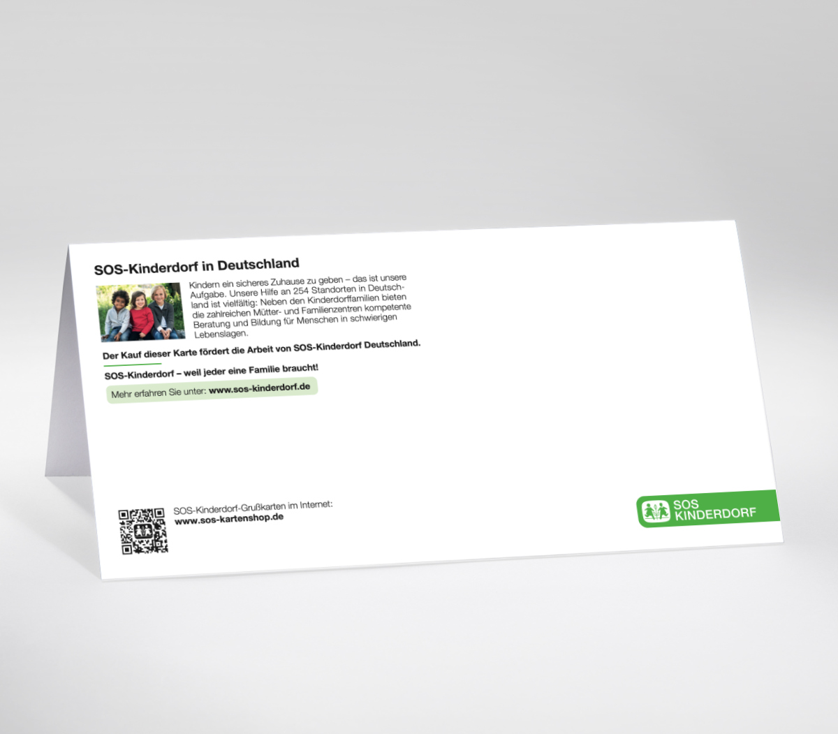 Rückseite einer weißen Grußkarte vom SOS-Kinderdorf. Oben links befindet sich ein Textblock mit der Überschrift „SOS-Kinderdorf in Deutschland“ in grüner Schrift. Darunter ein kurzer Infotext zur Arbeit des SOS-Kinderdorfs sowie ein kleines Farbfoto von Kindern. Im unteren Bereich ein grüner Hinweistext: Der Kauf dieser Karte fördert die Arbeit von SOS-Kinderdorf Deutschland. Rechts unten ist das grüne SOS-Kinderdorf-Logo mit dem Hinweis auf den Kartenshop www.sos-kartenshop.de zu sehen.