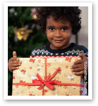 Kind mit lockigem Haar hält ein weihnachtlich verpacktes Geschenk vor einem geschmückten Weihnachtsbaum und lächelt in die Kamera